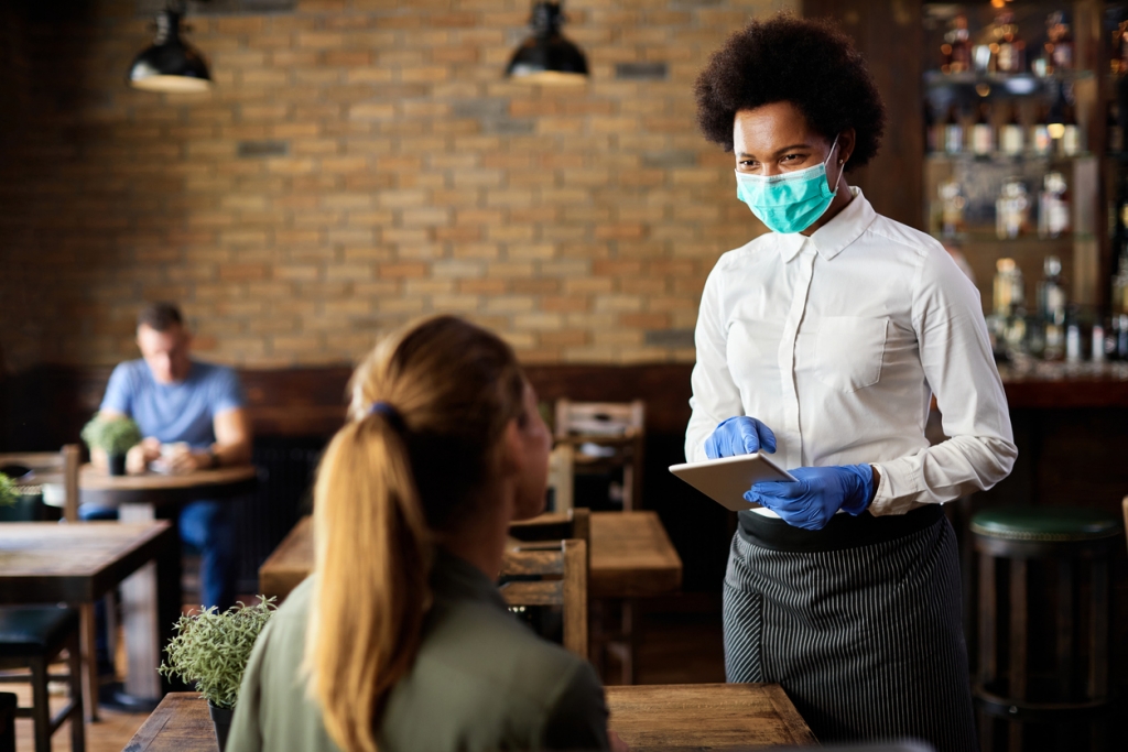 Happy black waitress wearing protective face mask while taking order from customer on a touchpad in a cafe.